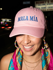 Woman wearing a pink embroidered dad cap reading “Mala Mía” during a nighttime outing.