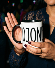 Woman holding a white mug with black interior and handle featuring “Cojones” in tall black lettering at night.
